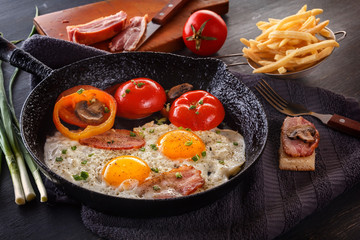 Fried eggs with bacon and tomatoes on an old cast-iron pan with french fries on a gray table. Close-up