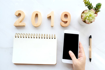 2019 wood letter, blank notebook paper and hand holding smart phone with blank screen on white marble background, 2019 new year mock up, template, flat lay