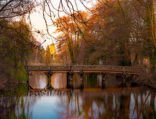 Bridge over a dark small lake with a red mill shining in the evening sun in the background .