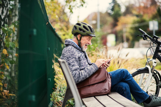 A senior man with electrobike sitting on a bench outdoors in town, using smartphone.
