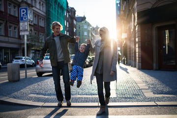 A small toddler boy with parents crossing a road outdoors in city at sunset.