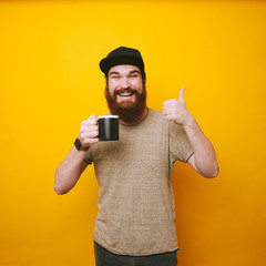 Happy bearded man holding black cup of coffee and showing thumbs up over yellow background