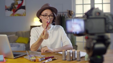 Medium shot of young woman in fedora hat and glasses recording makeup tutorial and explaining how to use products and brushes