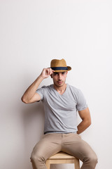 A confident hispanic young man with hat sitting on a stool in a studio.