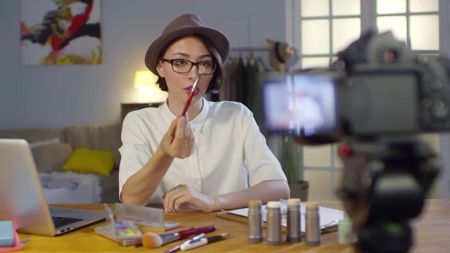 Medium shot of young woman in fedora hat and glasses recording makeup tutorial and explaining how to use products and brushes