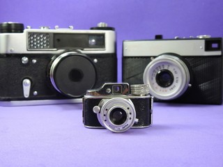 Photo cameras: in the foreground, the miniature of a vintage camera. Behind, two vintage blurred analog cameras. Lilac background.