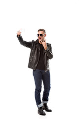 stylish adult man in leather jacket showing rock sign and taking selfie on smartphone isolated on white