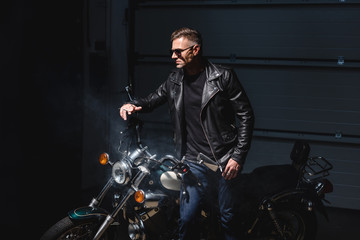 Fototapeta na wymiar fashionable guy in black sunglasses standing by motorcycle in garage