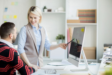 Cheerful confident pretty web designer with blond hair standing at table and pointing at computer monitor while presenting her project to manager in office