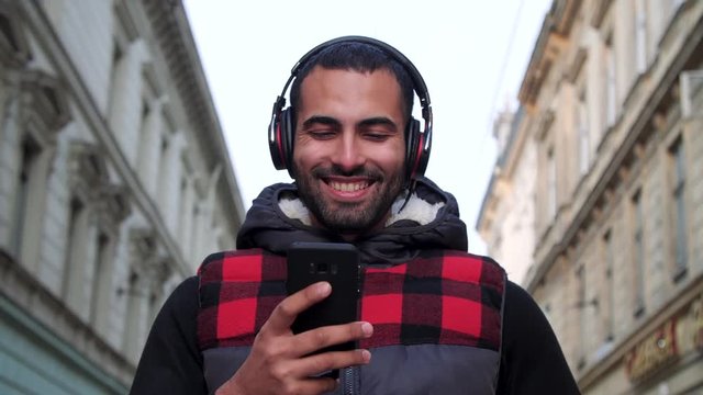Dancing and Smiling Attractive Guy Using his Smartphone for Texting Massage. He&rsquo;s Casual Dressed. Walking in Old European City Streets while Listening to Music. Headphones. Casual Lifestyle. People.