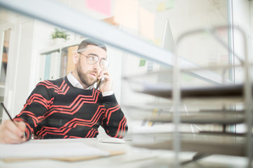 Serious frowning confident male manager in glasses sitting at table in modern office and talking by mobile phone while looking at computer screen