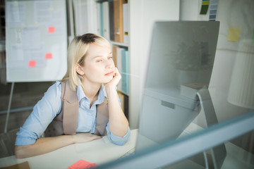 Serious thoughtful office woman sitting at table and leaning on hand while analyzing online data on computer in office