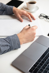 Businessman writing in notebook. White table with coffee laptop, glasses.
