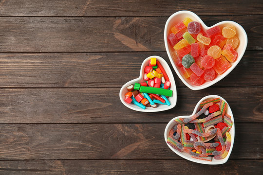 Flat lay composition with different yummy candies in heart shaped bowls and space for text on wooden background