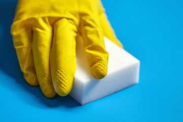 Human hand in yellow glove holds a white melamine sponge on blue background