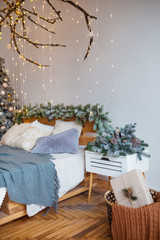 White cozy modern bedroom with holiday decoration. Wooden bed in scandinavian style room with festive Christmas tree in a pot and led garland lights. Home christmas decor.