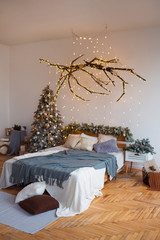 White cozy modern bedroom with holiday decoration. Wooden bed in scandinavian style room with festive Christmas tree in a pot and led garland lights. Home christmas decor.