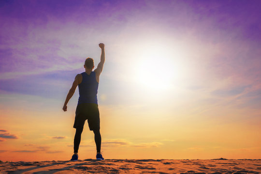Fit and healthy man standing with raised hand, silhouette against colorful sky with sunlight.