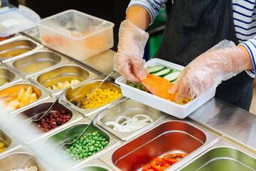 Fresh Salad bar counter with person's hands lifting salmon into a salad box for healthy and diet meal with smooth light and shadow.