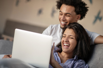 Cheerful happy African American young marriage laughing while watching movie on laptop computer at home interior.