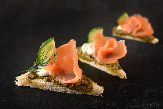 canapes in a row with smoked salmon, cucumber, pesto and dill garnish on dark stone with copy space, close up
