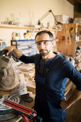 Waist up portrait of mature carpenter posing in workshop, standing next to electric disk saw