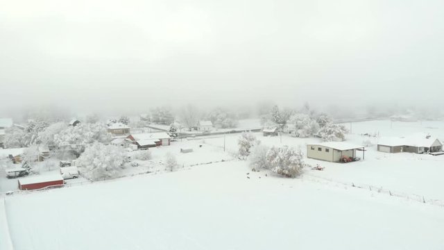 Aerial-Flying by snowy fields and a rural small town main street with snow covered buildings and trees and flying parallel to a dense fog bank