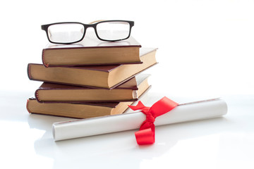 A parchment diploma scroll, rolled up with red ribbon beside a stack of books on white background.