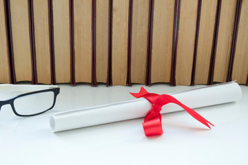 A parchment diploma scroll, rolled up with red ribbon beside a stack of books on white background.