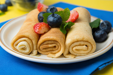 Rolled up crepes, pancakes on a plate with blueberries and strawberries. Yellow background.