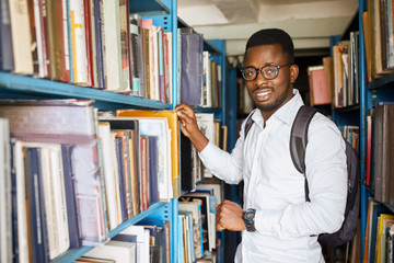 Young African-American man in glasses and white shirt standing between the bookshelves looking at...