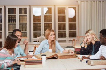 Fototapeta premium Mixed-race group of university students with a focus on blonde caucasian woman doing linguistic researches, turning over the pages of big old reference textbook at library.