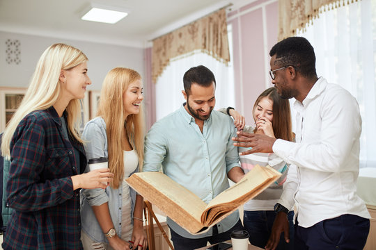 Handsome Arabian young bearded man surrounded by multiracial male and female students holding in hands big ancient book in library