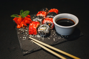 Sushi roll with cream cheese and fried salmon. Being picked up with chopsticks from soy sauce, with more rolls on black dish in the background. Soy sauce dripping.