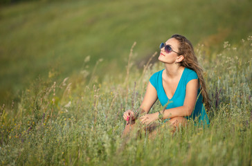 Naklejka na ściany i meble Young and beautiful caucasian girl with long hair in sunglasses sits in the meadow on a sunny day. Pretty woman is sitting on the grass and resting.