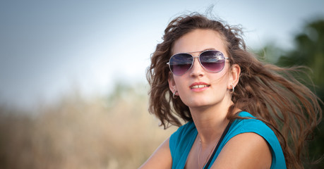 Young and beautiful caucasian girl with long hair in sunglasses sits in the meadow on a sunny day. Pretty woman is sitting on the grass and resting.