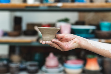 Master class concept. Cropped close up photo of workmanship lady in her workwear she stand against blurred wooden shelf background indoor workspace hold small ceramics clay tea cup on hand