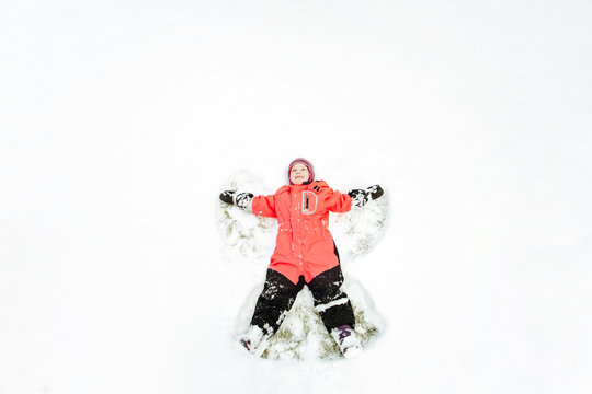 A child in a pink winter jumpsuit having fun, lying on the snow, arms and legs apart. Top view