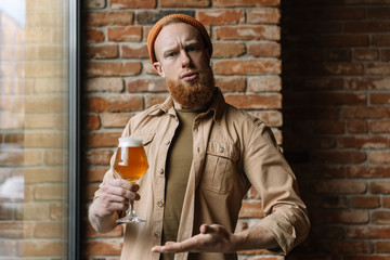 Bearded hipster man with funny emotional face holding glass of beer, drinking alcohol in pub....