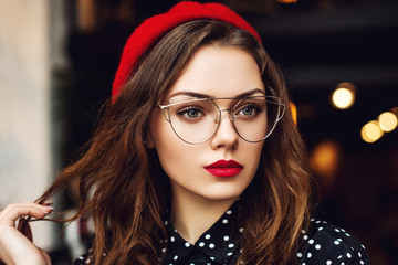 Close up portrait of young beautiful fashionable woman with red lips makeup, long hair, wearing stylish transparent glasses, red beret, polka dot blouse. Lights on background. 