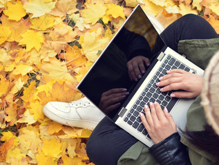 Girl in hipster with laptop in autumn park. A woman in a cap using a laptop while sitting on fallen leaves. Freelancer in the hat uses remote communication technology. Remote work. View from above