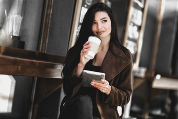 Beautiful girl uses a tablet and drinks coffee, sitting in a cozy cafe.