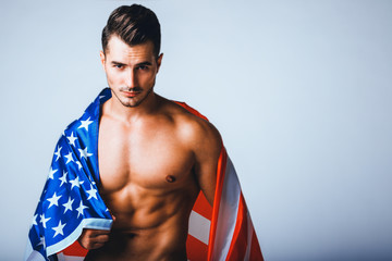 Portrait of shirtless handsome young man with stylish haircut posing with American flag over gray background. Perfect hair & skin. Close up. Studio shot