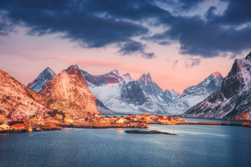Reine village and snow covered mountains at beautiful sunrise in winter. Lofoten islands, Norway. Landscape with blue sea, snowy rocks, houses and traditional rorbus, colorful sky with clouds. Coast