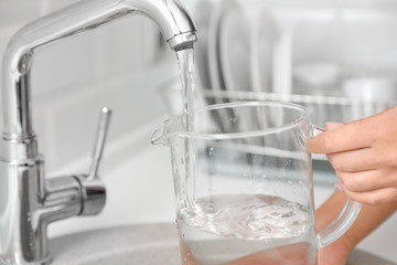 Woman pouring water into glass jug in kitchen, closeup