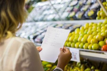 young woman holding shopping list in portuguese with fruits background in supermarket