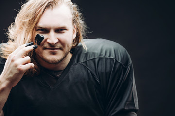 Portrait of confident fair-haired American football male player with war paint applied on his face looking at camera over black background