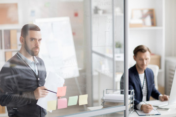 Portrait of thoughtful bearded man planning work sticking notes on glass wall in office, copy space
