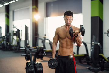 Fototapeta na wymiar Attractive fitness man with naked torso working out with dumbbells during training biceps in the gym. Sporty and healthy concept.
