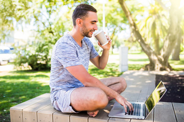 Portrait of young happy man with laptop and cup of coffee on bench in park